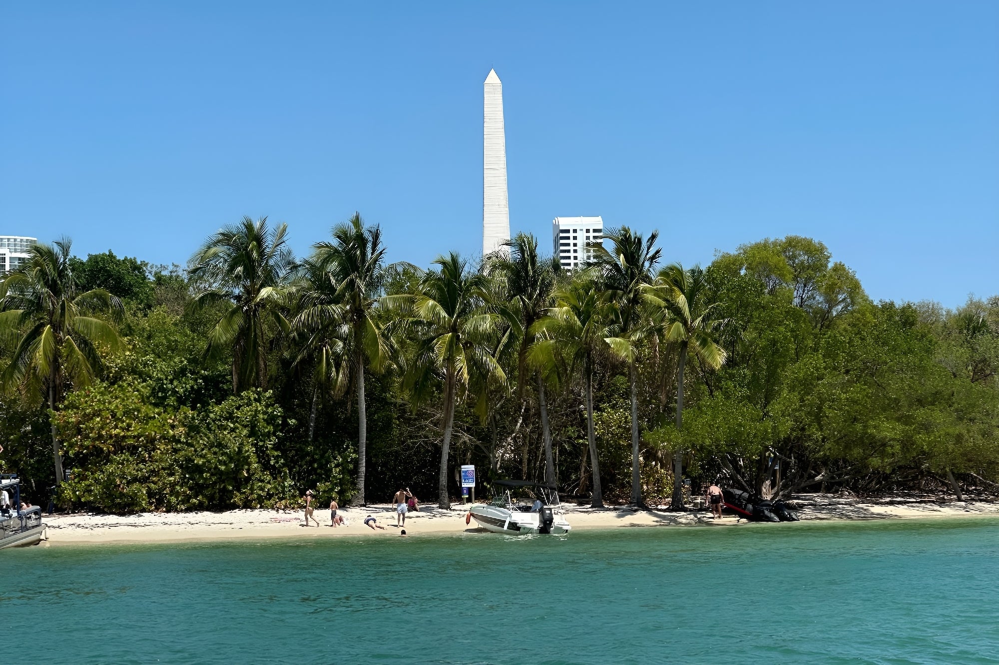 A sandy beach with palm trees borders clear blue water. Several people and boats are visible. A tall white obelisk towers in the background against a clear sky.