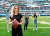 A woman holding a football stands on a field with three people behind her, two women and one man, in a stadium setting.