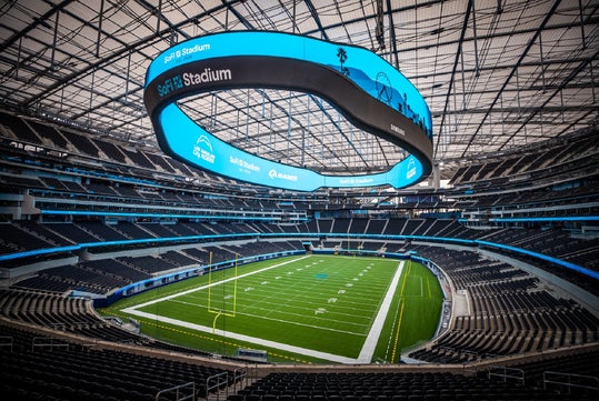 Empty football stadium with a large oval video board suspended above the field, displaying "SoFi Stadium" and various logos. Stadium seating surrounds the green turf field below.