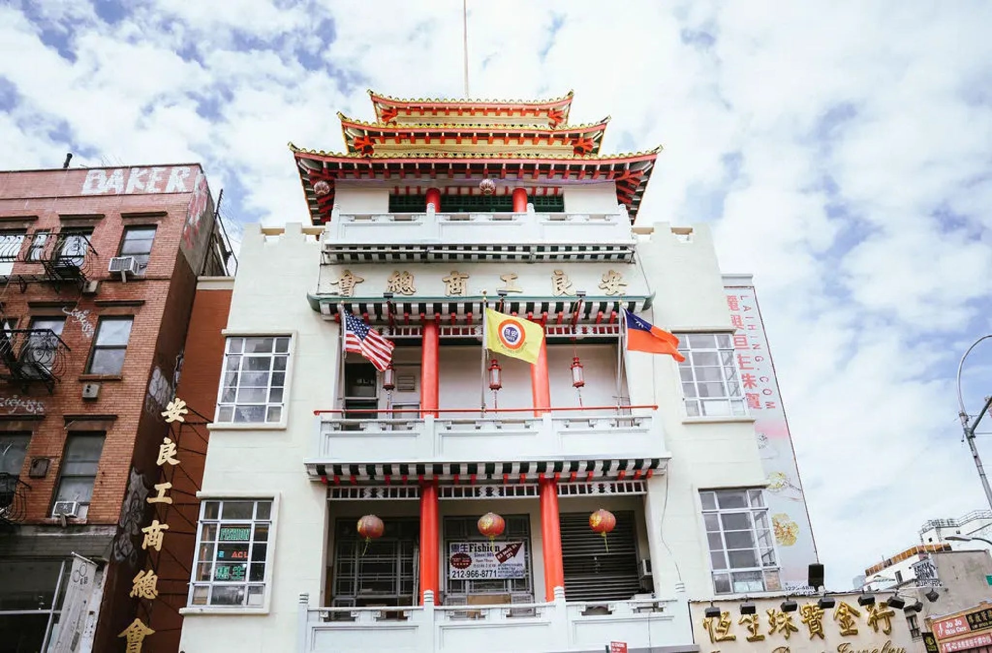 A multi-story Chinese-style building with red trim, hanging lanterns, and American and Chinese flags displayed on the balcony, set against a partly cloudy sky.