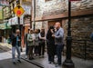 A group of people stand in line on a narrow city street, some eating food, with stone buildings and signs in the background.