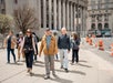 A group of people walk across a city sidewalk near large government buildings and traffic cones on a sunny day.