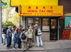A group of people stand in line outside Hing Tai Inc, a business with a yellow sign on Mosco Street in a city setting.