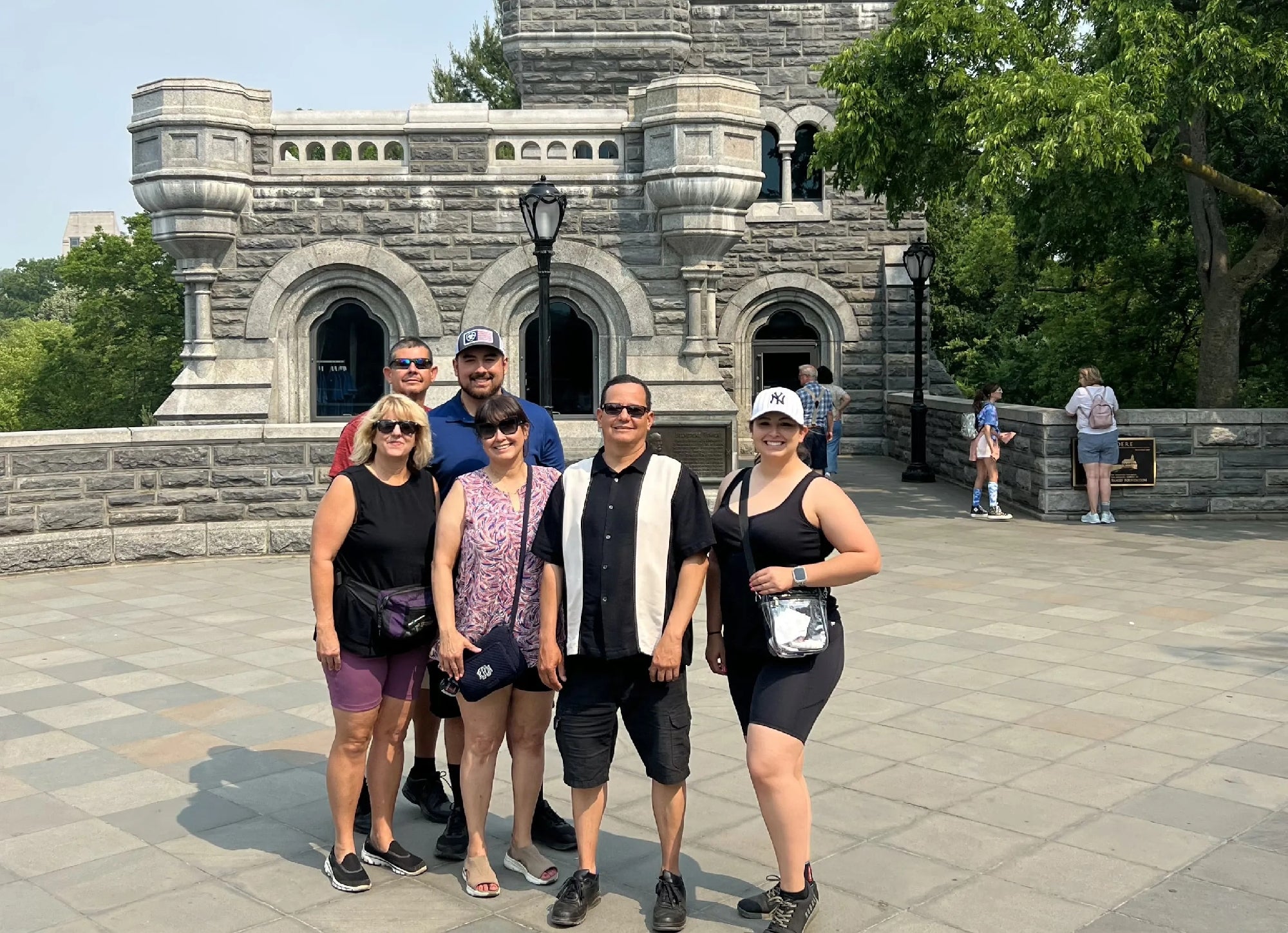Five adults pose for a group photo in front of a stone castle-like building on a sunny day; other people are visible in the background.