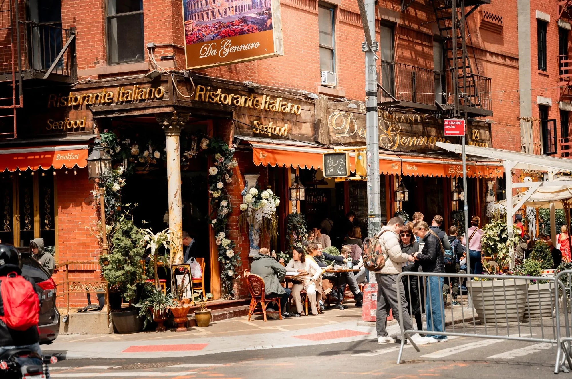 People dine outside a brick Italian restaurant on a city street corner, surrounded by plants and flowers, as pedestrians walk by and interact near the entrance.
