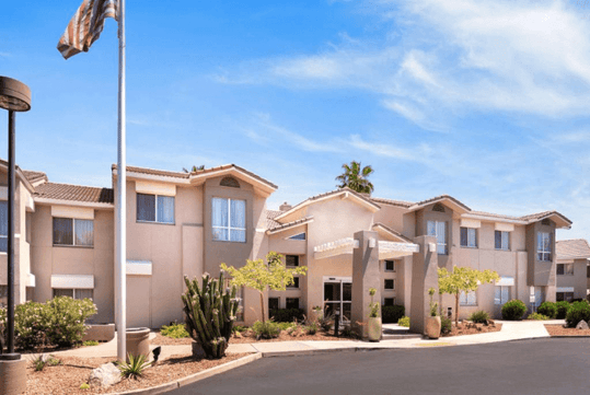 Two-story building with a beige exterior, surrounded by desert landscaping, under a clear blue sky. An American flag is visible on the left.