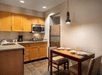 A small kitchen with wooden cabinets, stainless steel appliances, and a dining table set for two. The space is lit by overhead and pendant lights.