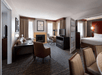 A spacious hotel room with a living area featuring a fireplace, desk, TV, and seating. To the right, there's an open door to a bedroom with a bed and another TV.