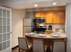 A kitchen with a stainless steel refrigerator, oven, and microwave. A countertop with four place settings and chairs is in the foreground. Light wooden cabinets are above the counter.