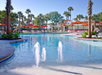 A resort pool with fountains in the foreground, surrounded by palm trees and lounge chairs with orange umbrellas.