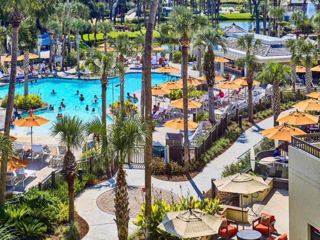 Aerial view of a resort pool area with people swimming and lounging under orange umbrellas surrounded by palm trees and greenery.
