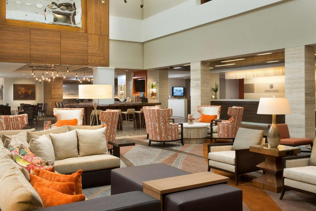 A modern hotel lobby with seating areas featuring beige and orange accents, contemporary chairs, and a decorative ceiling with pendant lights.