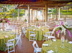 Outdoor event space with floral-decorated tables and white chairs on a wooden deck, overlooking a serene lake and garden. Draped pavilion with more seating in the background.