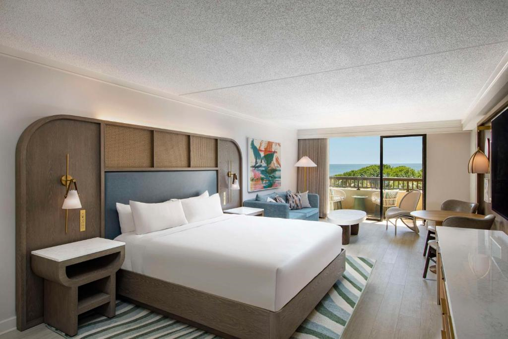 A spacious hotel room with a king-size bed, lounge area, and balcony view of greenery and the ocean. Modern decor includes a large painting, stylish lamps, and a striped rug.