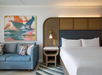 Modern hotel room with a large bed, blue sofa, wall art, and a small side table with a lamp.