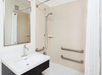 A bathroom with a white sink, faucet, and mirror on the left. A bathtub with a showerhead, grab bars, and a white curtain is on the right. Cream-colored tiles cover the wall.