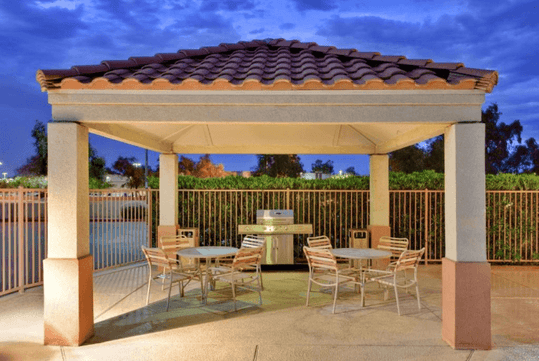 A gazebo with a tile roof features a barbecue grill and two sets of round tables with chairs, set on a paved surface, surrounded by a metal fence.
