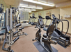A gym room with exercise equipment including treadmills, exercise bikes, and weight machines. A mirror covers one wall, and there are shelves with towels.