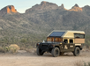 A rugged off-road vehicle with a camper topshell is parked on a dirt road, surrounded by a desert landscape and distant rocky hills during daylight.