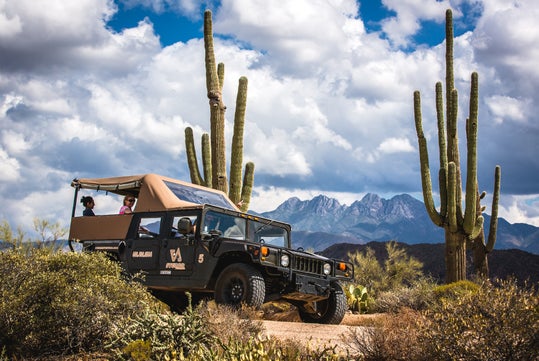 Off-road vehicle with canopy driving through a desert landscape with tall cacti and mountains in the background. Cloudy sky above.