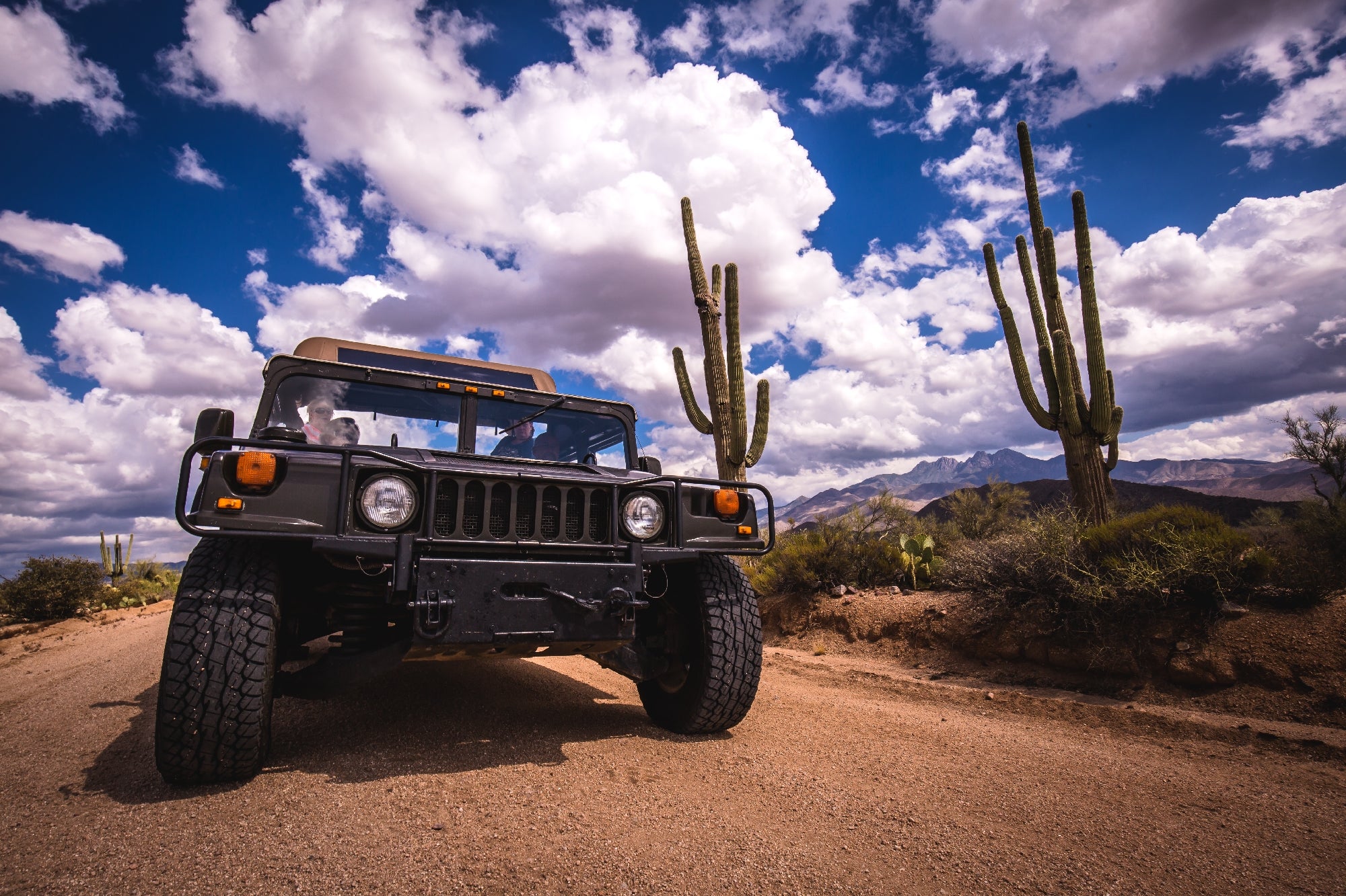 A large SUV travels on a dirt road in a desert landscape, surrounded by towering cacti under a partly cloudy sky.