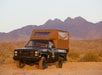 A black pickup truck with an attached brown camping tent drives on a desert terrain with rocky mountains in the background.