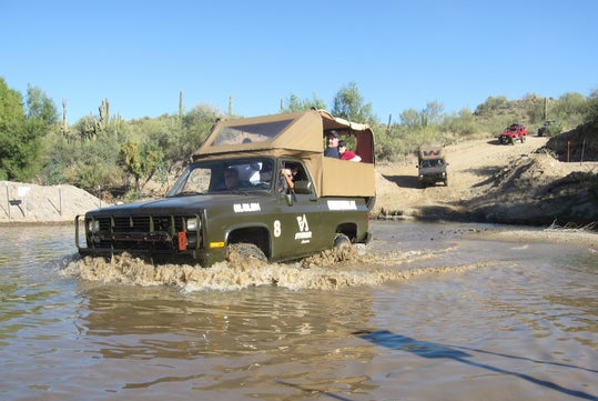 An off-road vehicle drives through a muddy water crossing in a desert landscape, with two people visible inside. Another vehicle follows in the background.