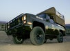 A modified black off-road truck with a roof tent is parked on desert terrain at dusk. The truck has a front grille guard and off-road tires.
