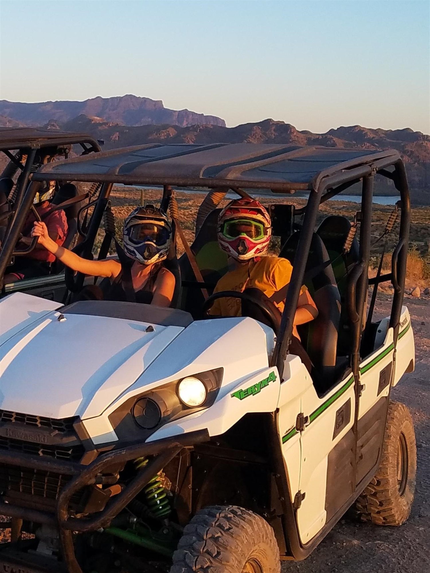 Two people wearing helmets sit in a white off-road vehicle on a dirt path, with mountains visible in the background.