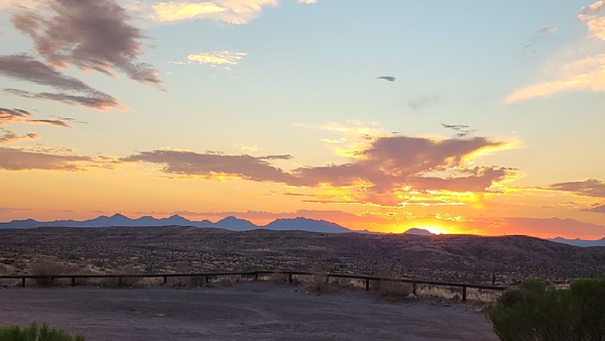 Sun setting over distant mountains, casting warm hues across the sky. Clouds partially cover the sun, with a flat landscape in the foreground.