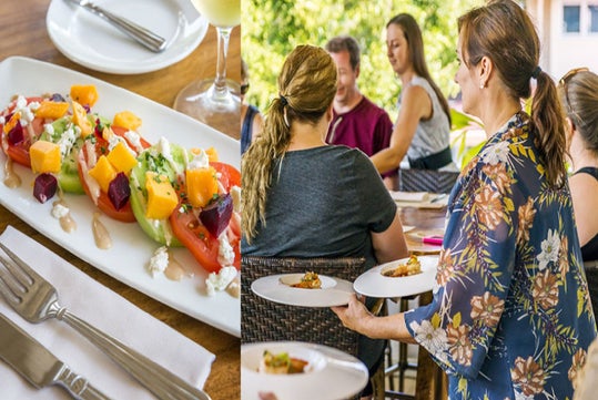 A server delivers plates to a group seated outdoors. A close-up shows a plated salad with beets, orange pieces, tomatoes, cucumber, and feta on the table beside a fork and knife.