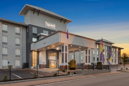 Modern hotel exterior at dusk, featuring a three-story building with a prominent entrance, flags, and a parking area.