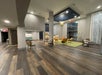 Spacious modern lobby with wooden flooring, chairs, and a green rug. Large windows and a high ceiling with a geometric light fixture.