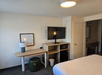 Hotel room interior with a bed, wall-mounted TV, desk with a lamp, green stool, small fridge, microwave, and framed art on a white wall. Door leading to the bathroom is partially visible.