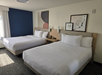 Hotel room with two neatly made double beds, a nightstand between them, and abstract artwork above. The room has a blue accent wall and gray carpet. Natural light enters through a window.