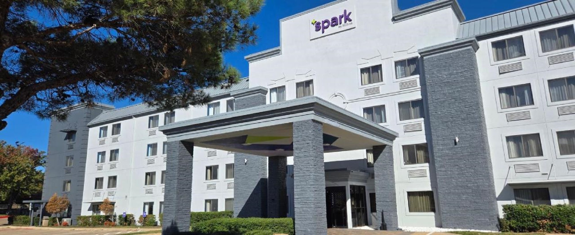 Exterior view of a four-story hotel, with a sign reading "spark," featuring a central entrance and surrounded by greenery.