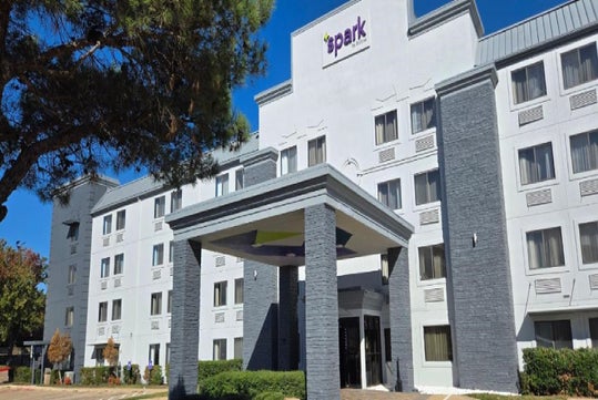 Exterior view of a four-story hotel, with a sign reading "spark," featuring a central entrance and surrounded by greenery.