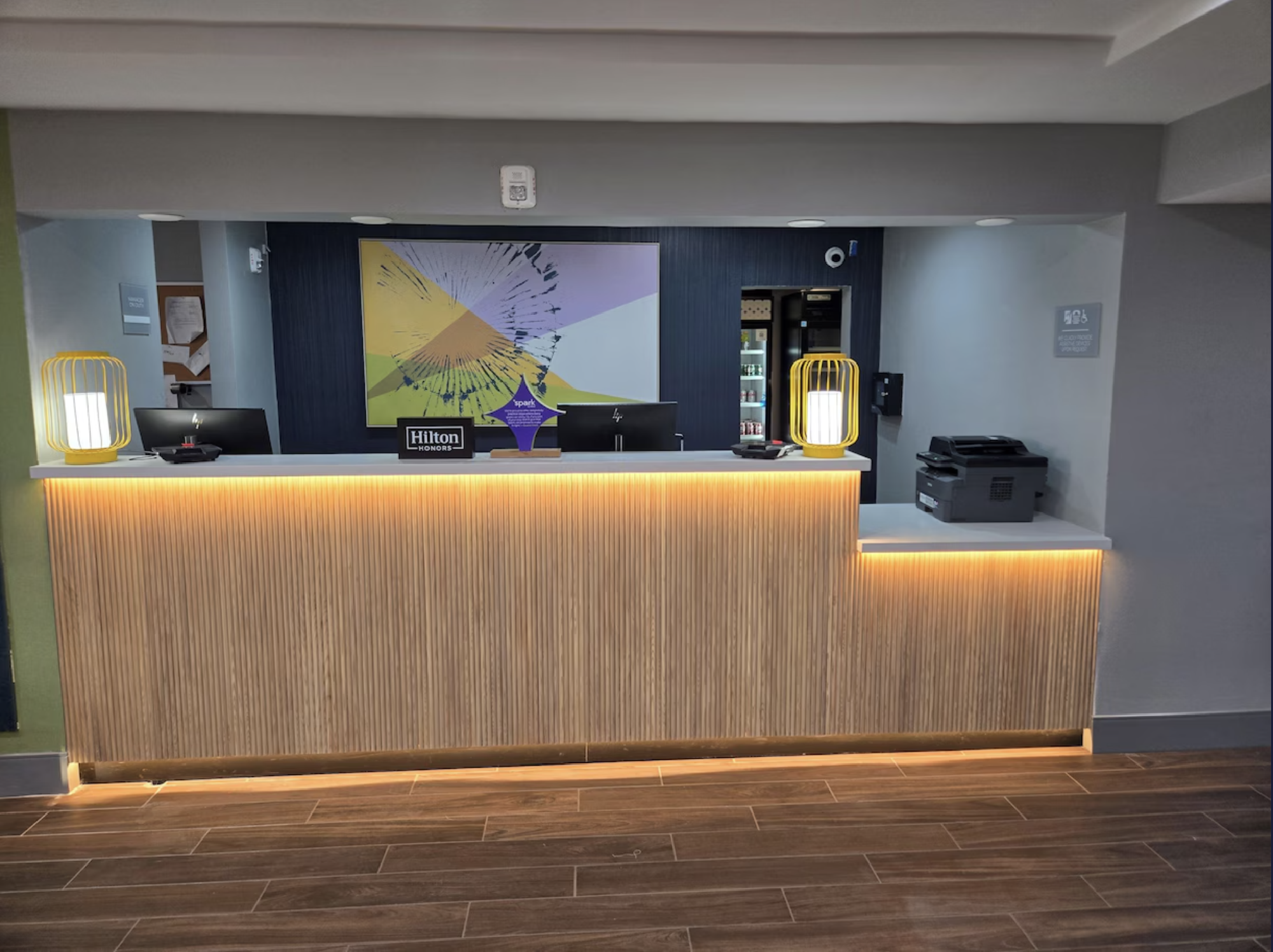 Hotel front desk with illuminated counter, abstract artwork on the wall, and two lamps.