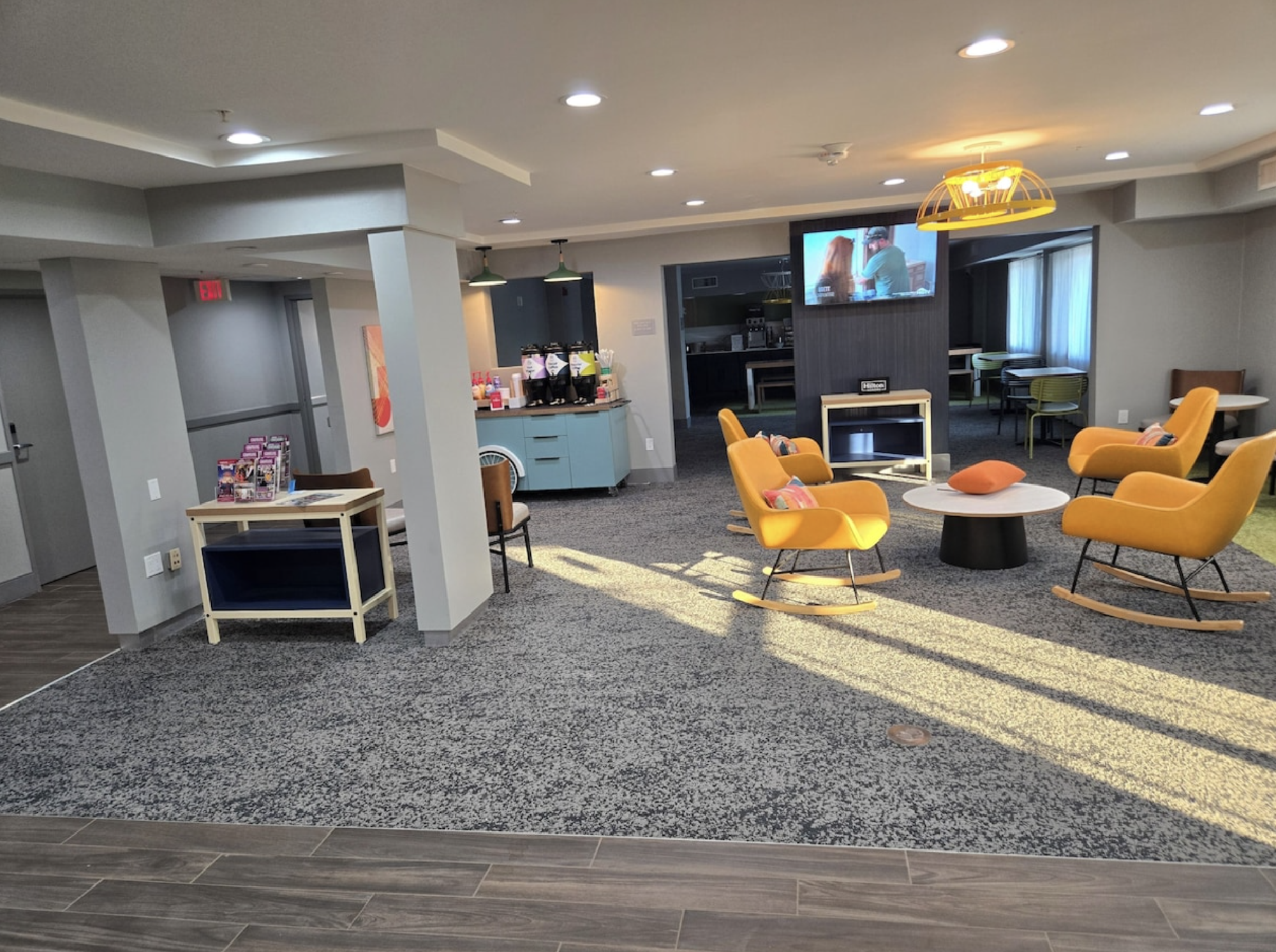 A modern lounge with yellow chairs, a round table, a wall-mounted TV, snack station, and a carpeted floor.