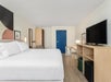 A modern hotel room with a king-sized bed, wall art, a TV on a wooden stand, a desk with a chair, and a blue door.