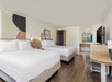 A hotel room with two double beds, a desk, chair, television, and a window with a view. The walls feature abstract art, and the floor is wooden.