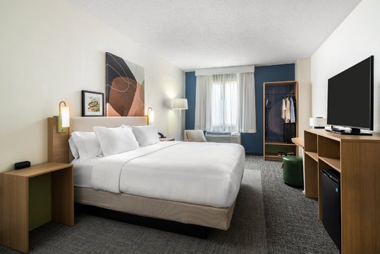 Hotel room with a neatly made king-size bed, abstract artwork above, a wall-mounted TV, open wardrobe with clothes, a window with sheer curtains, and a floor lamp.