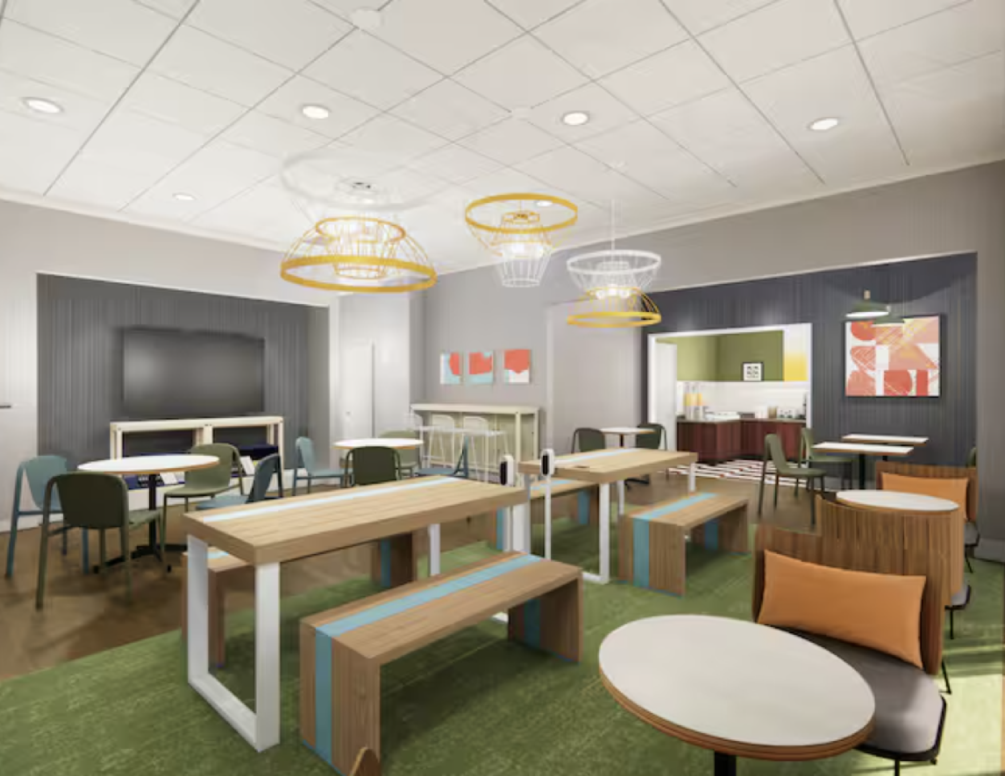 A modern communal space with wooden tables, chairs, and pendant lights. A TV is mounted on the wall, and a small kitchenette is visible in the background.