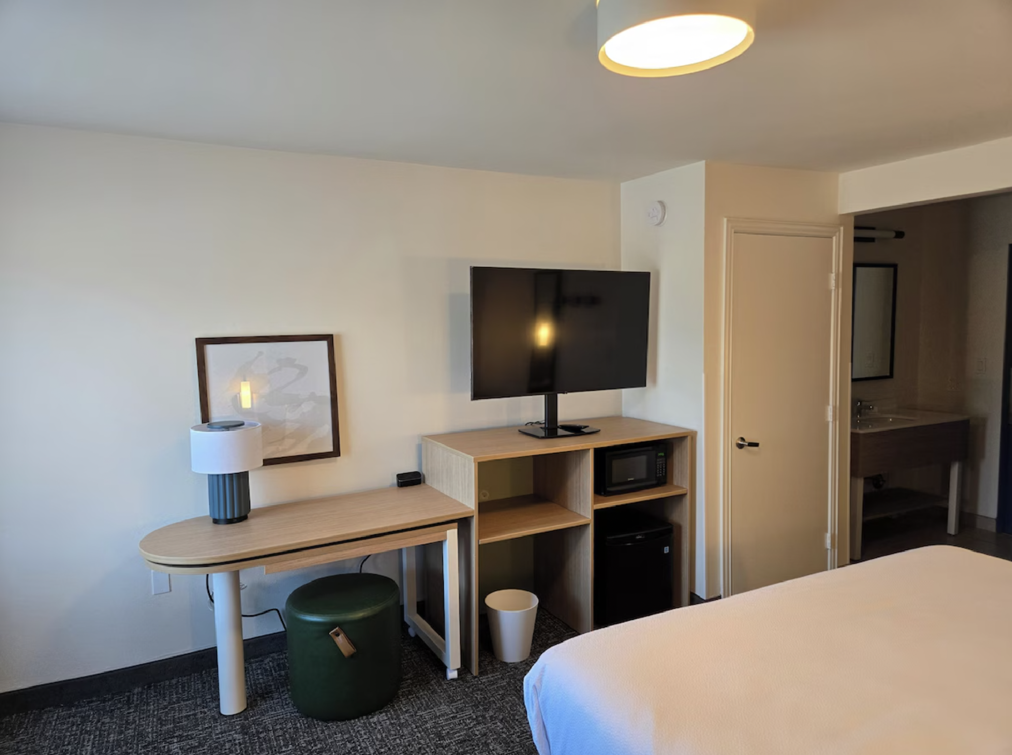 Hotel room with a wall-mounted TV, wooden desk with a lamp, small ottoman, mini fridge, and a glimpse of the bathroom.