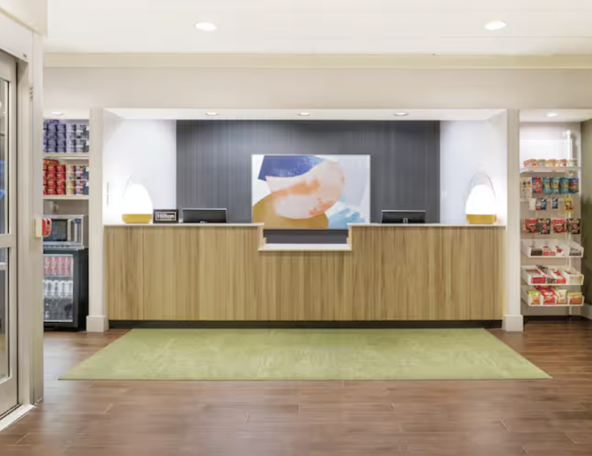 Hotel reception area with a wooden desk, abstract artwork, two lamps, and shelves containing snacks and beverages. A glass door is partially visible on the left.