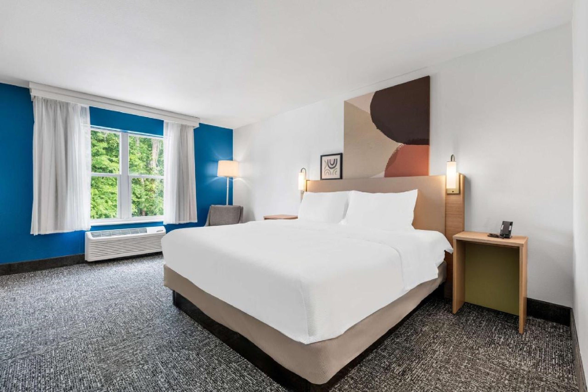 Hotel room with a large bed, white linens, modern artwork above the headboard, a window with white curtains, a floor lamp, and a nightstand with a phone.