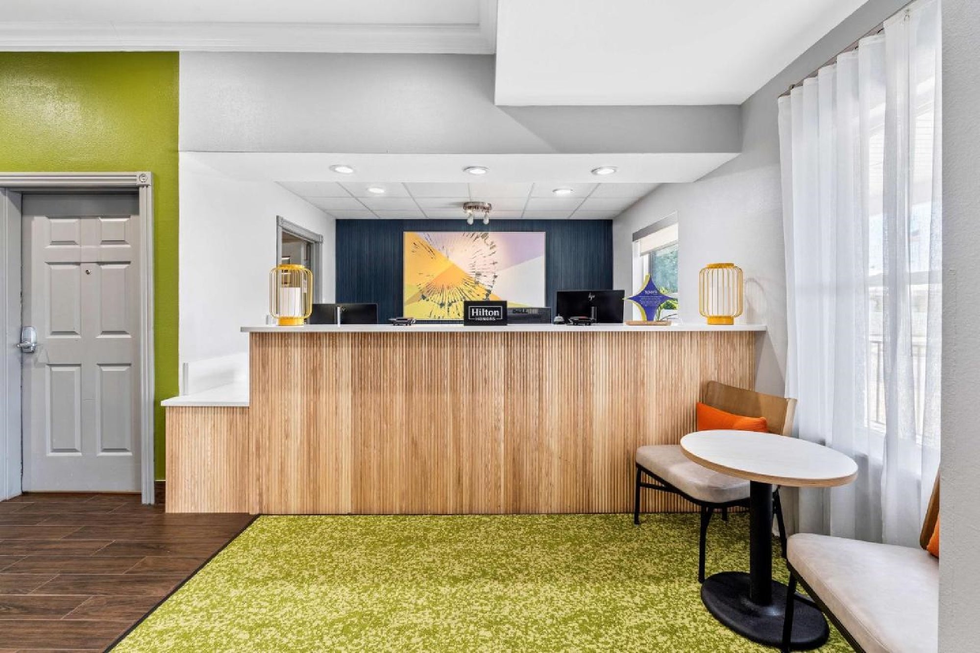 Hotel front desk area with wood and blue accents, a green rug, a round table with two chairs, and a colorful painting on the wall behind the desk.