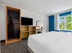 Hotel room with a large bed, a wall-mounted TV, a wooden desk, and an open closet with an iron and ironing board. A window shows greenery outside.