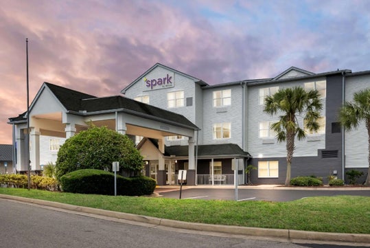 A three-story hotel with white siding and green roofing stands at sunset. The building is labeled "Spark" and features palm trees and a driveway in front.