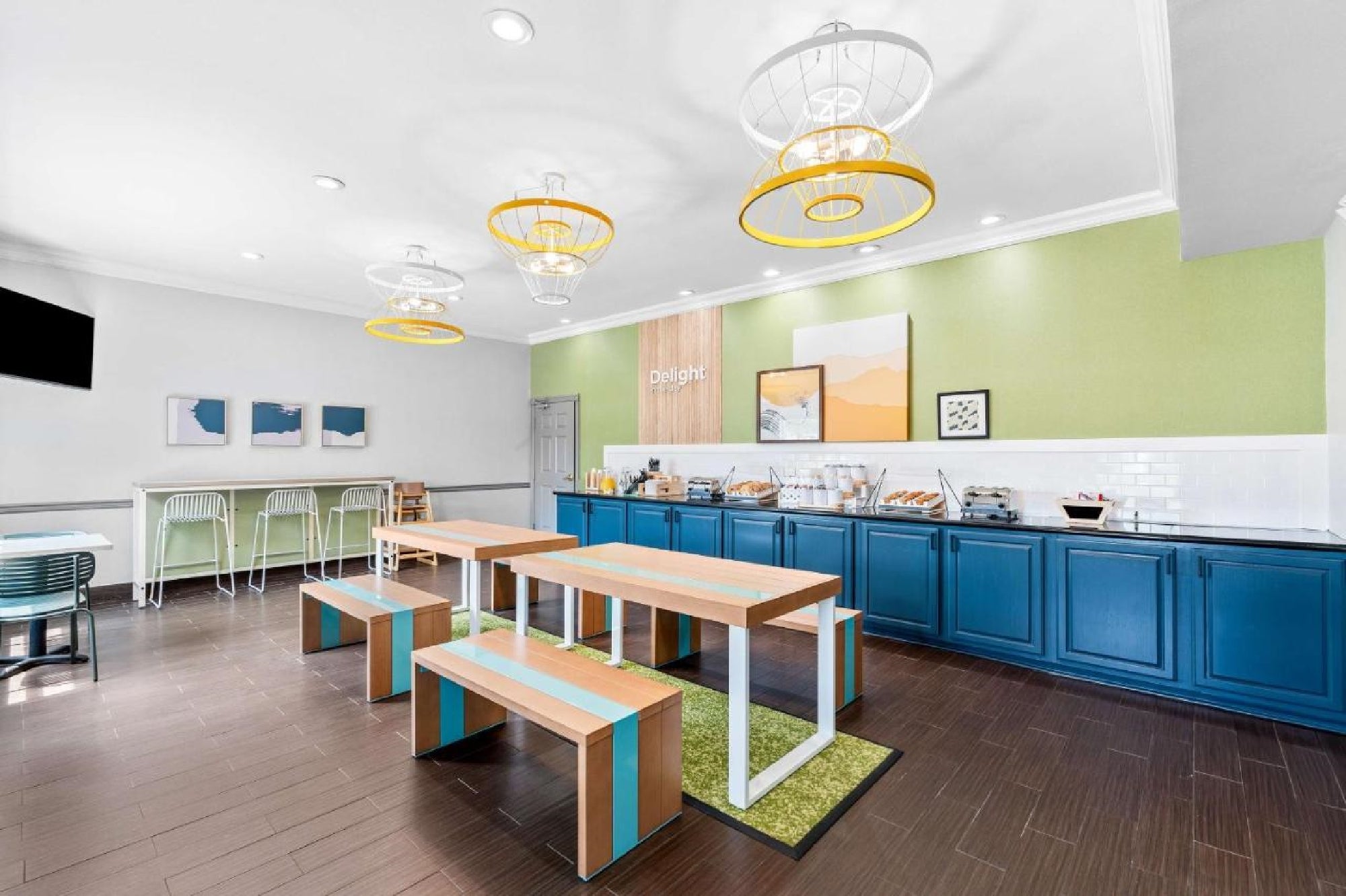 Bright dining area with tables and benches, blue cabinets, a buffet counter, green accent wall, and artistic wall decor. Overhead, round yellow and white lights are suspended from the ceiling.